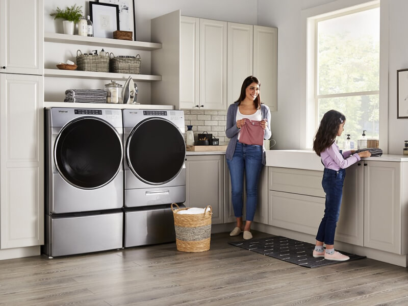 People folding clothes next to a Whirlpool® Washer and Dryer set in a modern laundry room