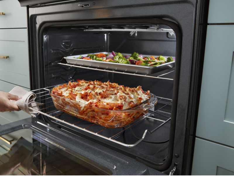 Person pulling out lasagna from the low oven rack while vegetables cook on the top rack