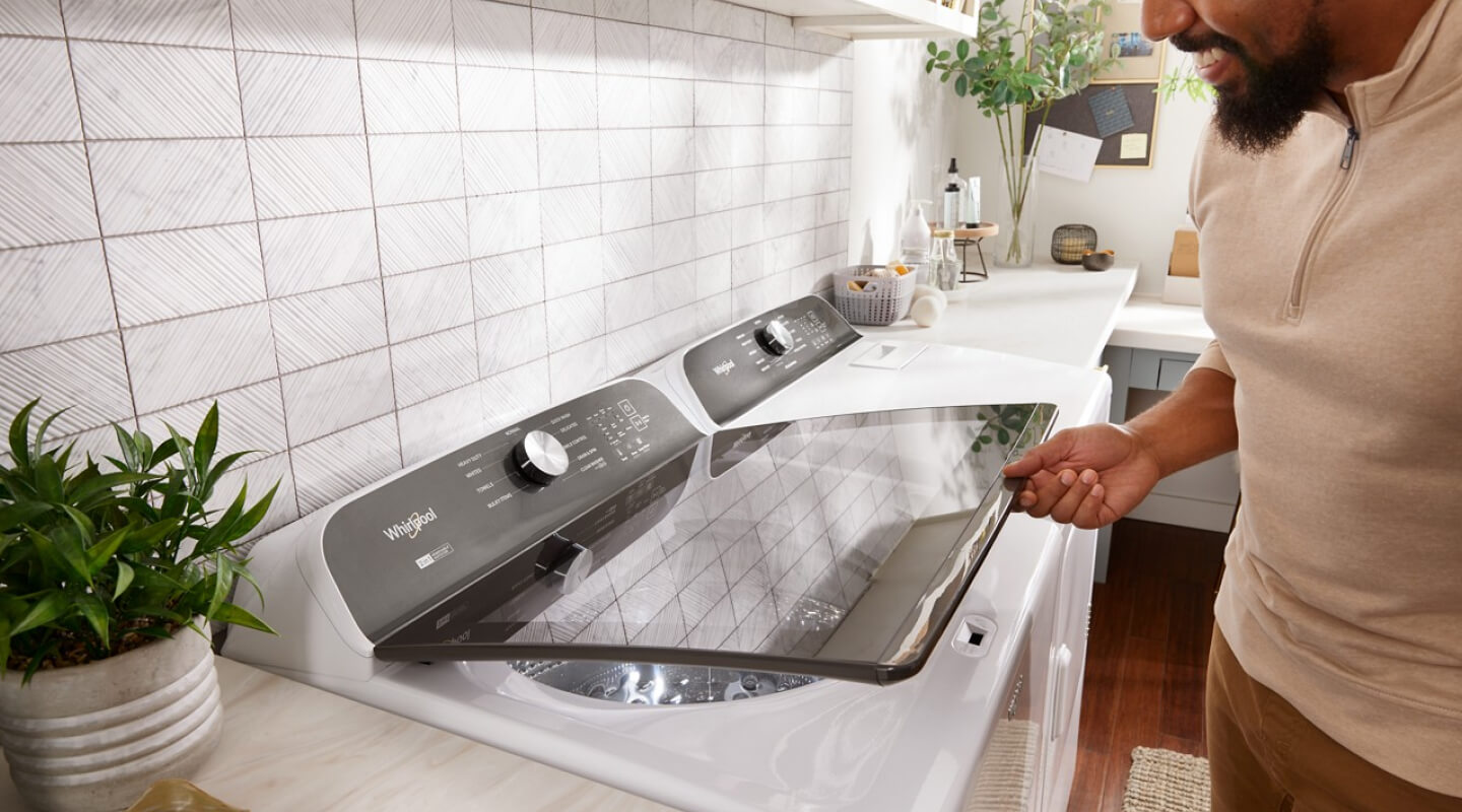 Person opening a white Whirlpool® Top Load Washer that is next to a white dryer in a laundry room