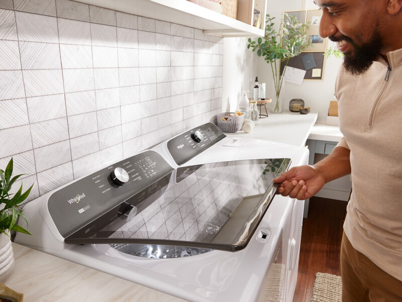 Person opening a white Whirlpool® Top Load Washer that is next to a white dryer in a laundry room