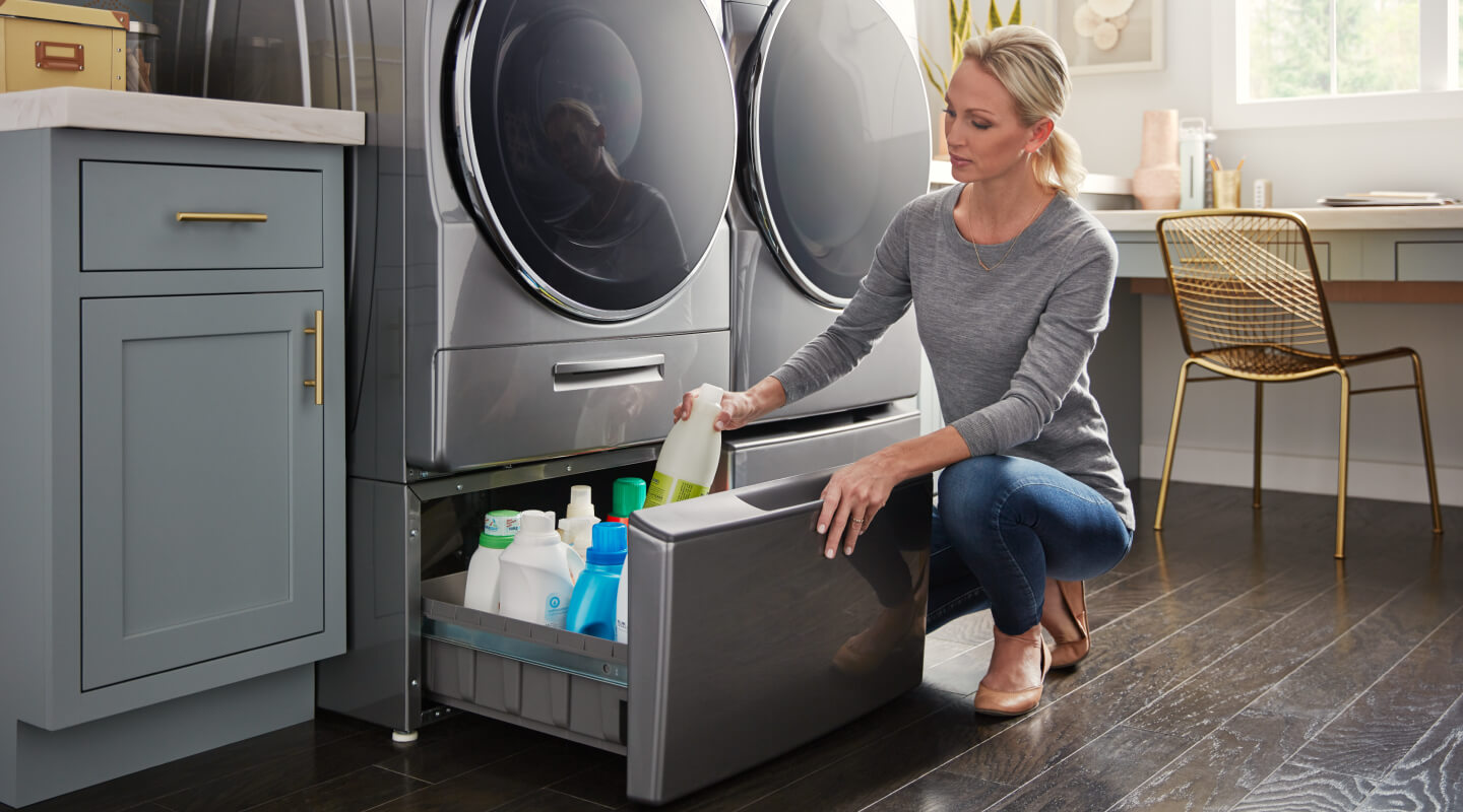 A person putting away detergent in a laundry pedestal that’s under a gray front load washer and next to a front load dryer