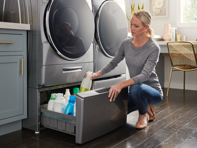 A person putting away detergent in a laundry pedestal that’s under a gray front load washer and next to a front load dryer