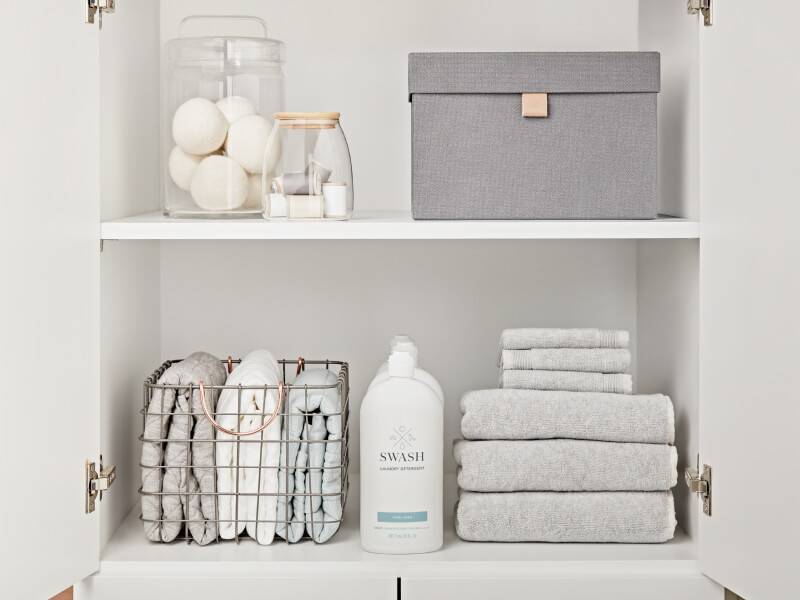 Swash® Laundry Detergent on the bottom shelf of a white cabinet between stacks of gray towels