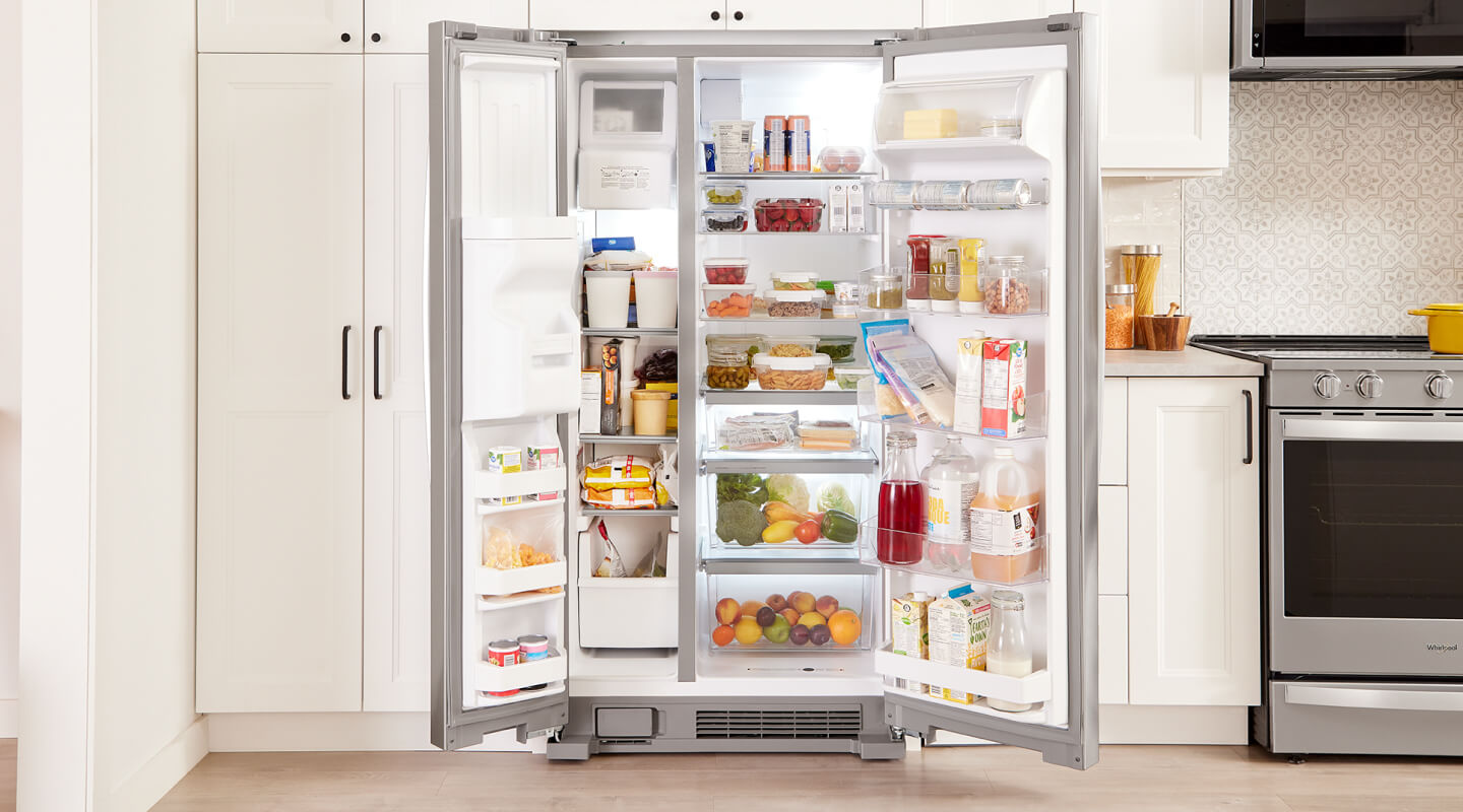An open, side-by-side fridge filled with food and surrounded by white cabinets