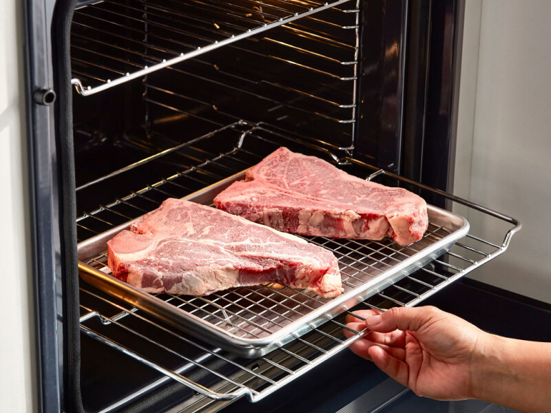 A person sliding a rack of raw steaks into an oven