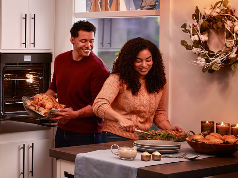 A couple preparing a dinner of roasted chicken, vegetables and bread rolls