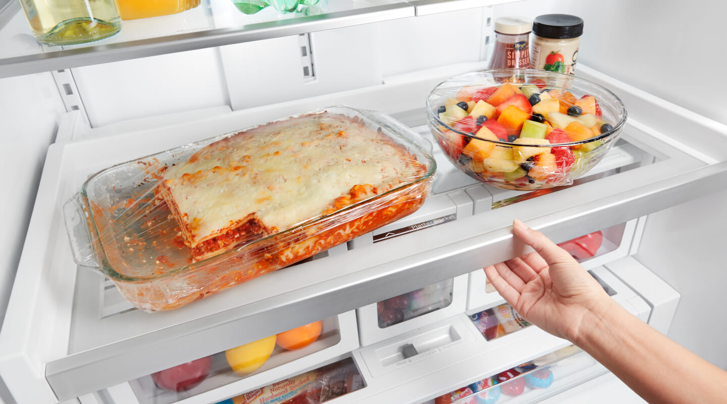 A lasagna and a fruit bowl sitting on a fridge shelf