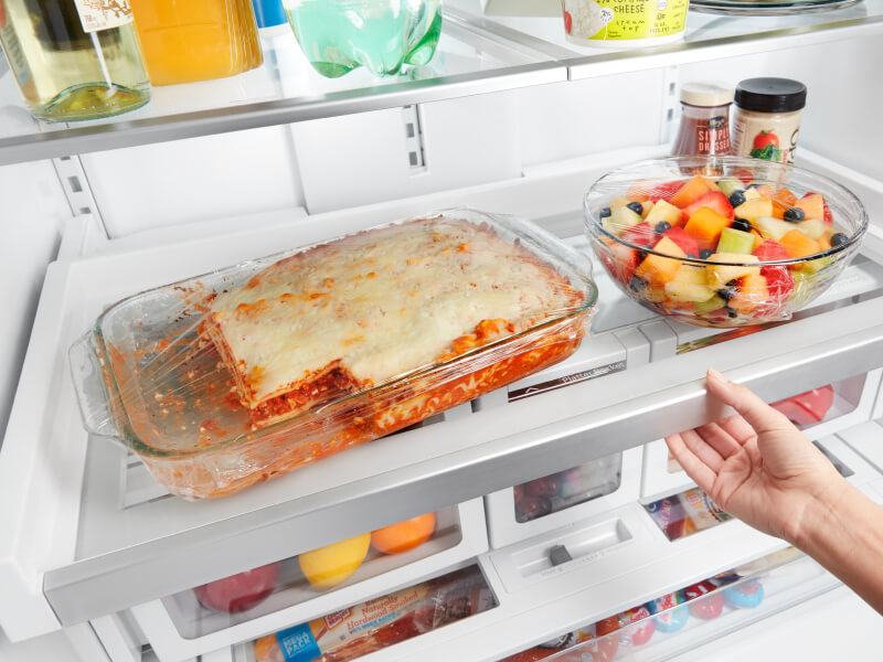 A lasagna and a fruit bowl sitting on a fridge shelf
