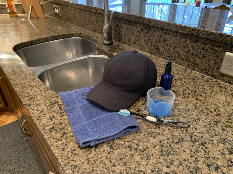 A black cloth baseball cap on a countertop with a toothbrush, towel and cleaning supplies