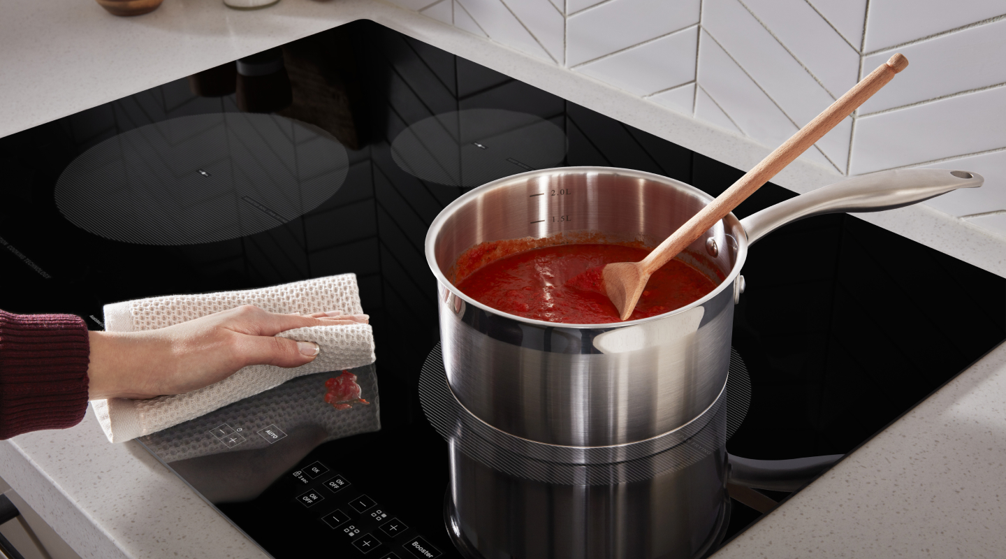 Person wiping sauce splatter off of induction cooktop next to sauce pan