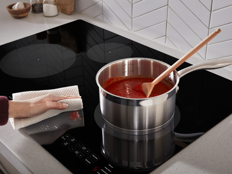 Person wiping sauce splatter off of induction cooktop next to sauce pan