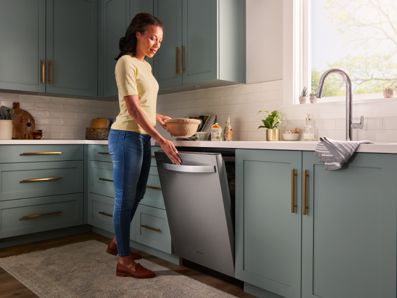 A person closing a stainless steel dishwasher surrounded by teal cabinetry.