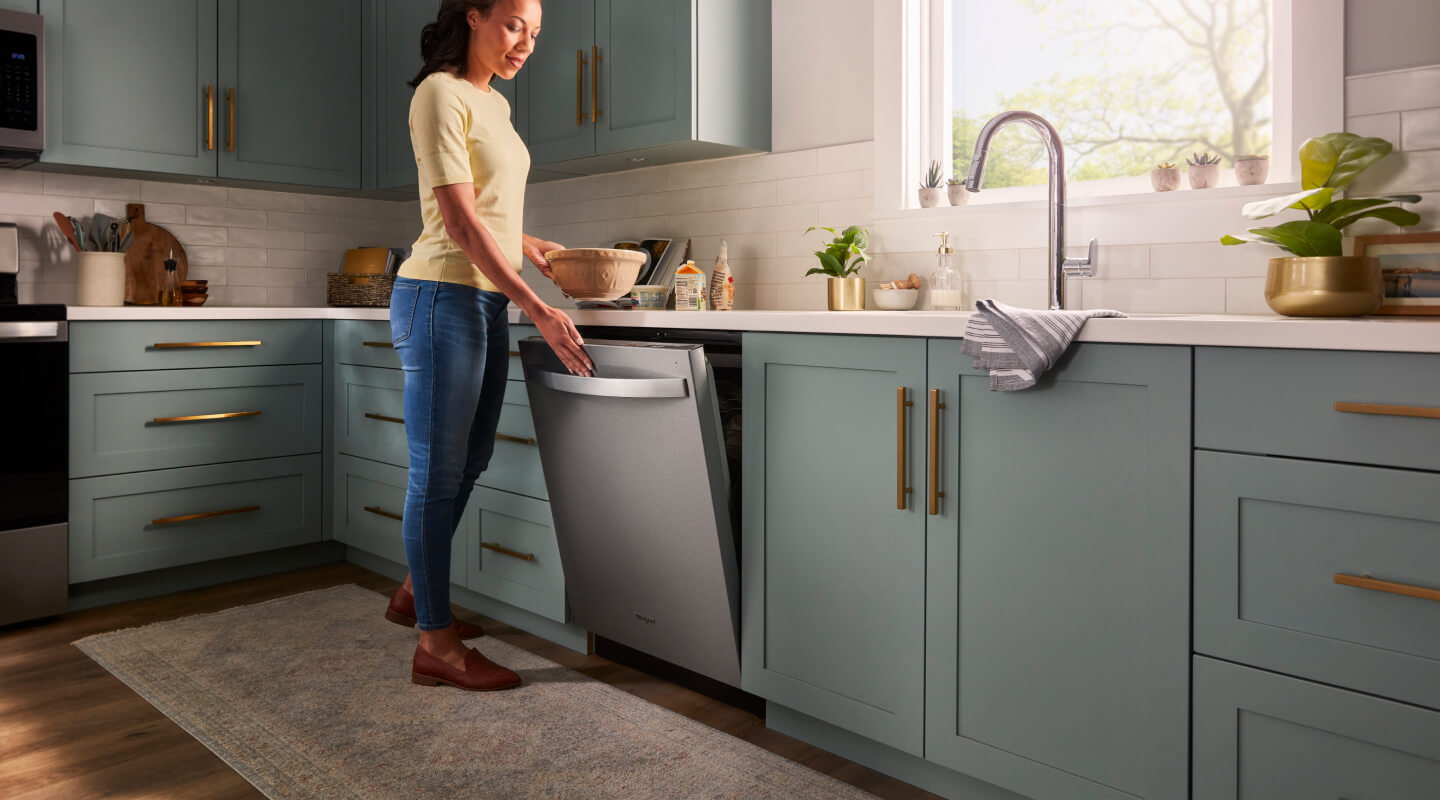 A person opening a Whirlpool® Built-In Dishwasher with Top Control and a towel handle in a kitchen with sea green cabinets and white countertops