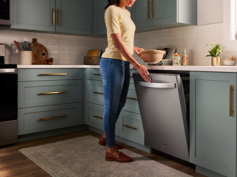 A person opening a Whirlpool® Built-In Dishwasher with Top Control and a towel handle in a kitchen with sea green cabinets and white countertops