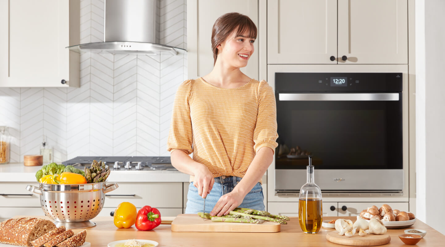 A person chopping asparagus on a wooden cutting board placed on a kitchen island with a cooktop in the background behind her