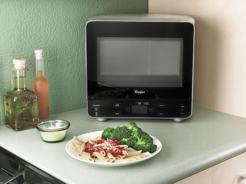 A Whirlpool® Countertop Microwave on a kitchen countertop next to a plate of pasta and broccoli.