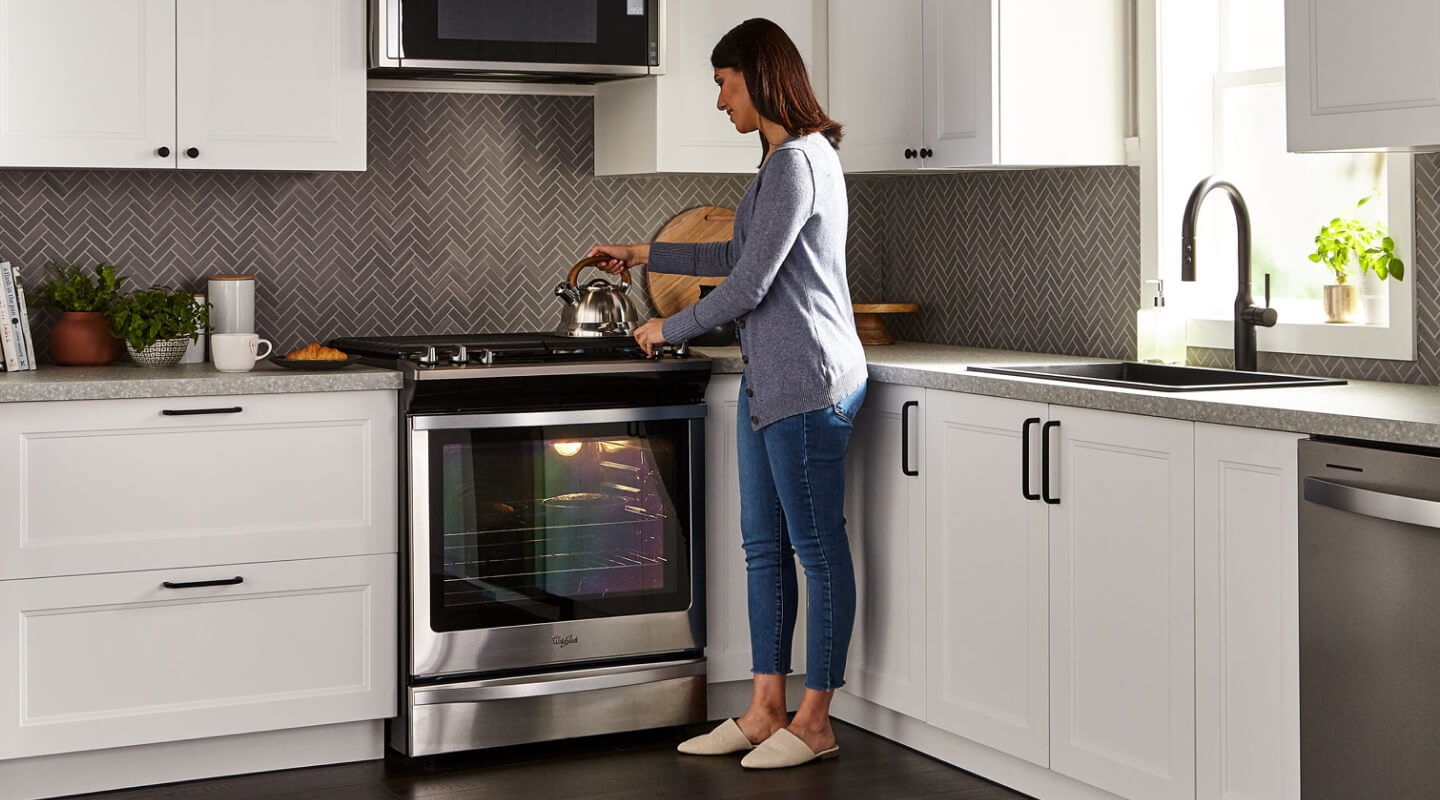 A person placing a tea kettle on the stovetop of a Whirlpool® gas oven in a modern kitchen.