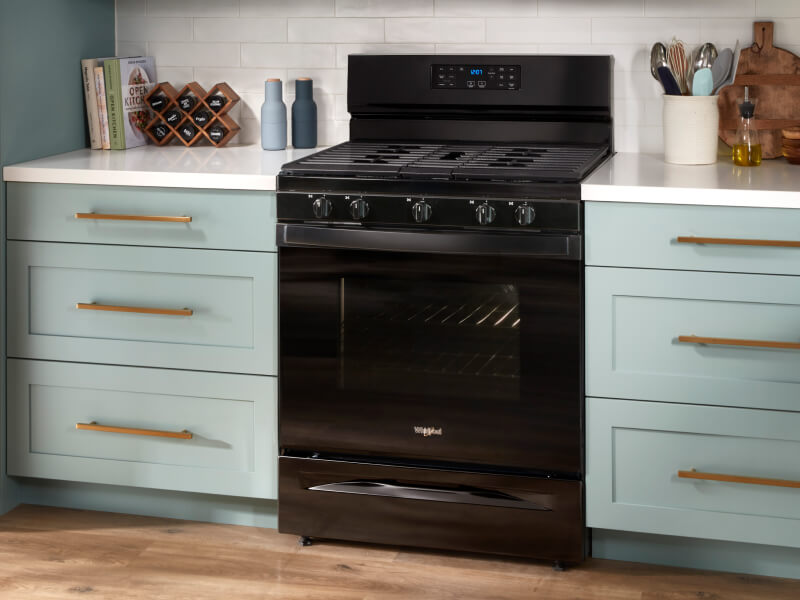 A black gas range between sea green cabinets with white countertops
