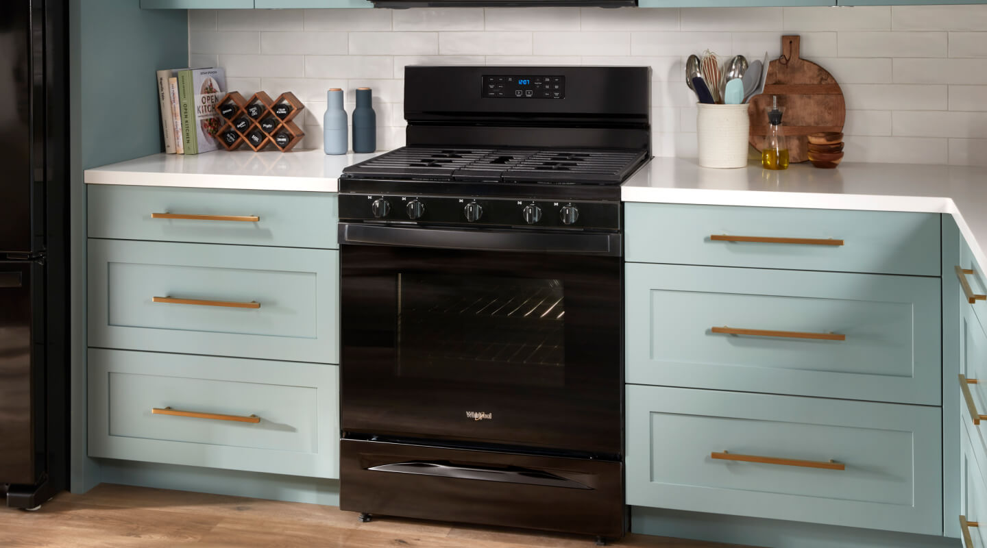 A black gas range between sea green cabinets with white countertops